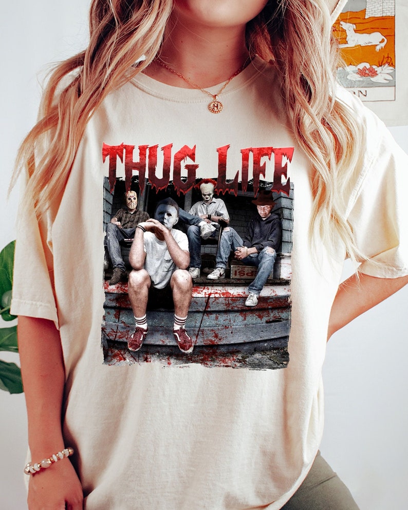 May include: A cream-coloured t-shirt with a graphic that reads "THUG LIFE" in dripping red letters. The graphic shows four horror film characters seated on a porch. The image has a distressed, vintage aesthetic.