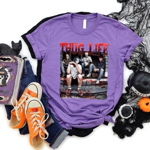 May include: A lilac t-shirt with the text "THUG LIFE" in red, featuring a graphic of four people on a porch. The image is surrounded by Halloween decorations, including a black cat figurine, a decorative book, and orange trainers.