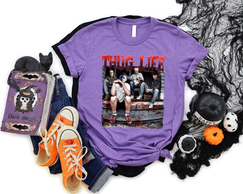 May include: A lilac t-shirt with the text "THUG LIFE" in red, featuring a graphic of four people on a porch. The image is surrounded by Halloween decorations, including a black cat figurine, a decorative book, and orange trainers.