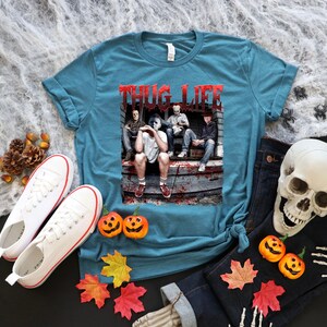 May include: Teal t-shirt with the text "THUG LIFE" in red lettering. The shirt features a graphic of horror movie characters. The image is surrounded by Halloween decorations, including pumpkins, a skull, and autumn leaves.