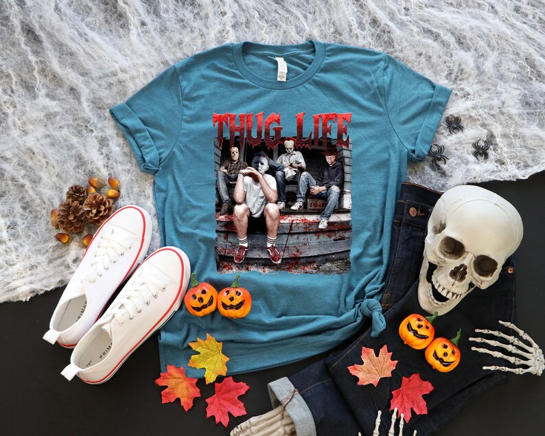 May include: Teal t-shirt with the text "THUG LIFE" in red lettering. The shirt features a graphic of horror movie characters. The image is surrounded by Halloween decorations, including pumpkins, a skull, and autumn leaves.
