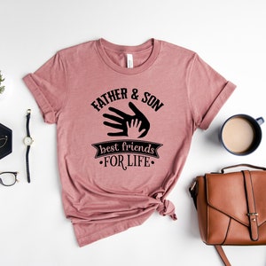 May include: A dusty rose t-shirt with the text "FATHER & SON best friends FOR LIFE" in black. The design features a hand-in-hand graphic. The shirt is styled with a watch, glasses, and a brown purse.