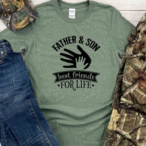 May include: Green t-shirt with the text "FATHER & SON best friends FOR LIFE" in black. The shirt is paired with blue jeans, a camouflage hat, and a camouflage jacket.