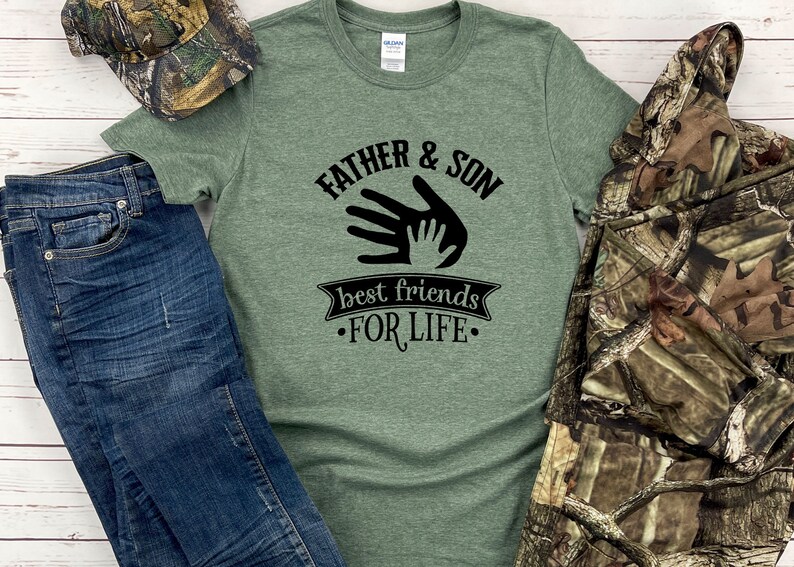 May include: Green t-shirt with the text "FATHER & SON best friends FOR LIFE" in black. The shirt is paired with blue jeans, a camouflage hat, and a camouflage jacket.