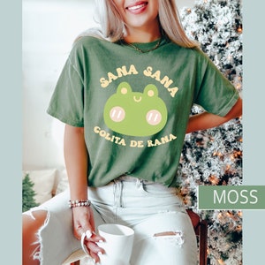 Puede incluir: Una camiseta verde con una rana de dibujos animados y el texto "Sana Sana Colita de Rana" en letras amarillas y blancas.