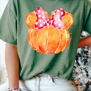 Op de afbeelding: Olijfgroen t-shirt met een aquarelontwerp van een oranje pompoen met een roze strik met stippen. Het woord "MOSS" wordt weergegeven in een groene rechthoek. Het t-shirt wordt door een persoon gedragen.
