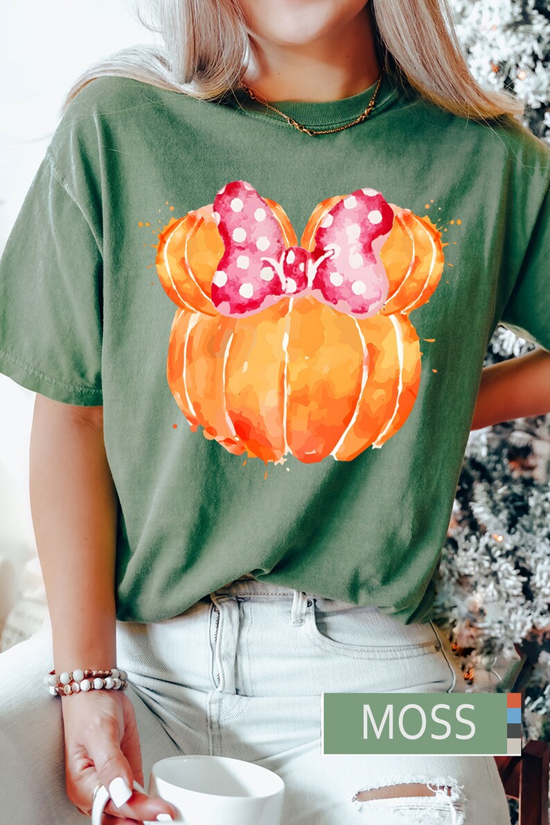 Op de afbeelding: Olijfgroen t-shirt met een aquarelontwerp van een oranje pompoen met een roze strik met stippen. Het woord "MOSS" wordt weergegeven in een groene rechthoek. Het t-shirt wordt door een persoon gedragen.