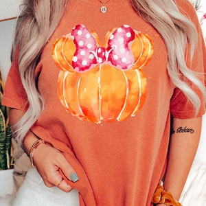 Op de afbeelding: Oranje T-shirt met een aquarel pompoen ontwerp en een roze strik met stippen. Het woord "YAM" wordt onderaan in een rechthoek weergegeven. Het shirt heeft een effen oranje kleur.