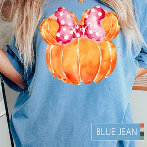 Op de afbeelding: Lichtblauw T-shirt met een aquarelontwerp van een oranje pompoen met een roze strik met stippen. Op het shirt staat "BLUE JEAN".
