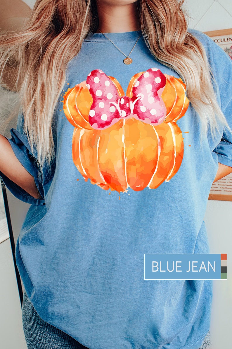 Op de afbeelding: Lichtblauw T-shirt met een aquarelontwerp van een oranje pompoen met een roze strik met stippen. Op het shirt staat "BLUE JEAN".