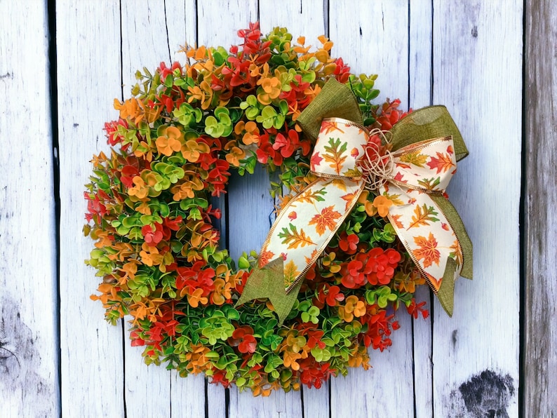 May include: A green, orange, and red fall wreath with a white and orange ribbon with a fall leaf pattern. The wreath is made of artificial leaves and is hanging on a white wooden door.