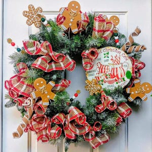 May include: A festive Christmas wreath with faux evergreen branches, red and white plaid ribbon, and gingerbread man and snowflake ornaments. A decorative plate with the text "Hot Christmas Cafe" is in the center.