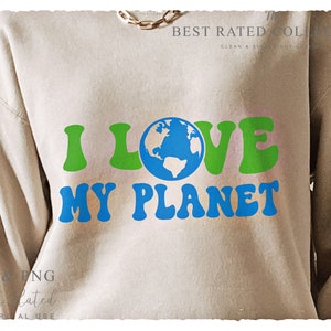 Puede incluir: Sudadera beige con el texto "I LOVE MY PLANET" en verde y azul. La palabra "LOVE" presenta un gráfico de un globo terráqueo. La sudadera también tiene el texto "SVG & PNG" y "Best Rated" en la parte inferior.