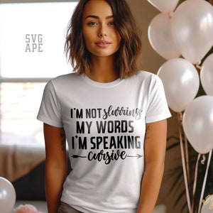 Può includere: T-shirt bianca con la scritta nera "I'M NOT slurring MY WORDS I'M SPEAKING cursive". La maglietta è a girocollo. L'immagine include anche palloncini bianchi e una persona.