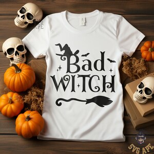 May include: White t-shirt with the words "Bad Witch" in black, featuring a witch on a broom, bats, and stars. The shirt is surrounded by pumpkins, skulls, and decorative elements, creating a Halloween theme.