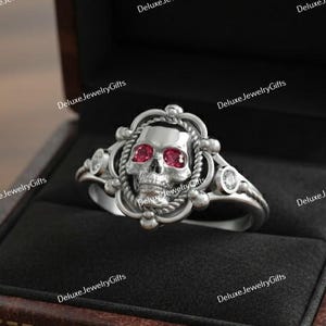 Puede incluir: Un anillo de calavera plateado con ojos de piedras preciosas rojas, exhibido en un joyero de cuero marrón. El anillo presenta detalles intrincados alrededor de la calavera y en la banda, con gemas claras adicionales.