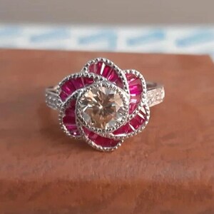May include: A silver ring with a large, round, champagne-colored diamond surrounded by a halo of smaller diamonds and a cluster of red rubies in the shape of a flower.