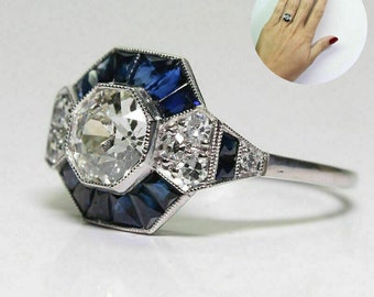 Op de afbeelding: Een zilveren ring met een grote, achthoekige middelste steen omringd door kleinere blauwe en witte stenen.
