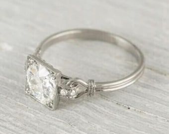 May include: A silver engagement ring with a square-shaped diamond set in a square frame. The ring has a delicate band with a twisted design and small diamonds on the side.
