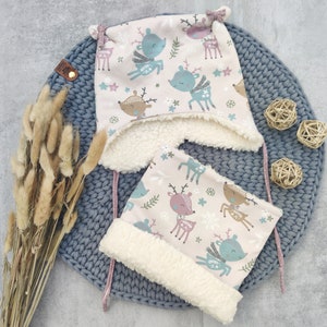 Könnte beinhalten: Zwei rosa und weiße Wintermützen mit einem Rentier-Muster. Die Mützen haben ein weißes Fleece-Futter und sind mit rosa und weißen Schnüren gebunden.