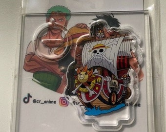 May include: Clear acrylic keychain featuring a colorful illustration of the Thousand Sunny ship from the anime series One Piece. The keychain also includes images of anime characters. The keychain is in a clear plastic sleeve.