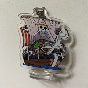 May include: A clear acrylic stand with a colorful illustration of a ship with a skull and crossbones on the sail. The ship is being pulled by a white creature with a ram's head. The stand is on a white background.
