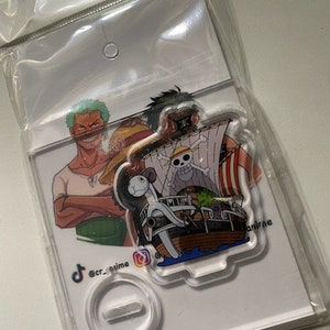 May include: A clear acrylic stand with a printed image of three characters from the anime One Piece. The characters are Luffy, Zoro, and Sanji. The stand is on a white background with the text "@cr_anime" and a TikTok logo.