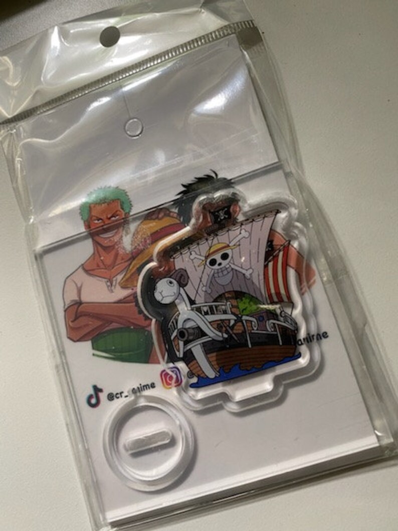 May include: A clear acrylic stand with a printed image of three characters from the anime One Piece. The characters are Luffy, Zoro, and Sanji. The stand is on a white background with the text "@cr_anime" and a TikTok logo.