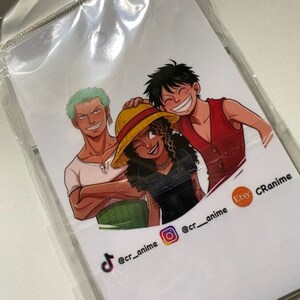 May include: A white card with a colorful illustration of three anime characters. The characters are smiling and standing together. The card has social media handles for @cr_anime on TikTok, Instagram, and Etsy.