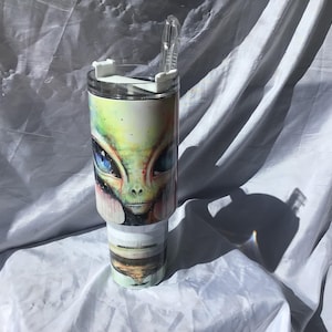 May include: A stainless steel tumbler featuring a detailed alien face design with blue eyes, green skin, and a cosmic background. The tumbler has a white lid with a clear straw and is approximately 10 inches tall.