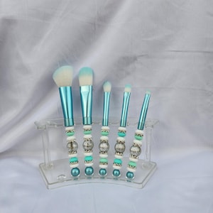 May include: A collection of five make-up brushes with light blue handles and white bristles. The handles are adorned with silver, white, and turquoise beads. The brushes are presented in a clear acrylic holder against a white backdrop.