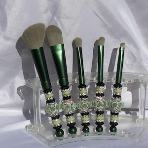 May include: A collection of five makeup brushes. Each brush has a green handle and light-coloured bristles. The handles are adorned with black and pale green beads, rhinestones, and tiny green rose embellishments. The brushes are in a clear acrylic holder.