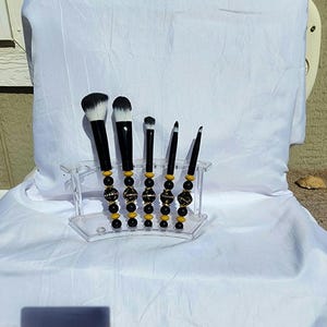 May include: A collection of five black and white makeup brushes with decorative handles. The handles feature black, gold, and yellow beads. The brushes are presented in a clear acrylic holder, set against a white backdrop.