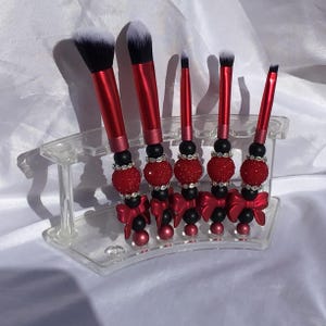 May include: A set of six makeup brushes with red handles and white bristles. The handles are decorated with black and red beads, rhinestones, and red bows. The brushes are displayed in a clear acrylic holder.