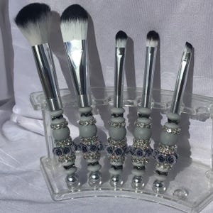 May include: A set of five makeup brushes with silver metal handles and white bristles. The handles are decorated with grey beads, rhinestones, and small dark blue roses. The brushes are displayed in a clear plastic holder.