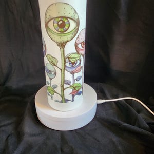 May include: White insulated tumbler with a colorful floral design featuring stylized eyes. The artwork includes green, blue, and red elements. The tumbler is on a white charging base.