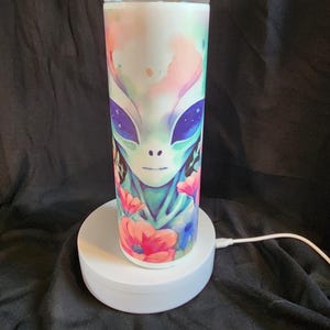 May include: A white stainless steel tumbler featuring a colorful alien face design with large purple eyes and floral accents. The tumbler has a clear lid and is displayed on a white rotating stand.