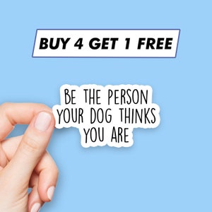 May include: White sticker with the text "BE THE PERSON YOUR DOG THINKS YOU ARE" on a light blue background. A hand is holding the sticker. The text "BUY 4 GET 1 FREE" is visible in the background.