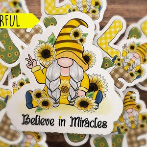 May include: A colorful sticker featuring a gnome with a yellow and black striped hat, holding a peace sign. The gnome is surrounded by sunflowers and the text "Believe in Miracles".