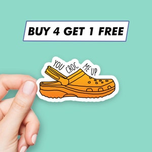 Puede incluir: Una ilustración de dibujos animados amarilla y naranja de un zueco con el texto "You Croc Me Up" en el lateral. La imagen es una pegatina con un borde blanco. El texto "BUY 4 GET 1 FREE" está en la parte superior de la imagen.
