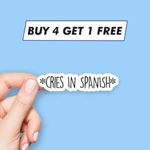 May include: White sticker with the text "*CRIES IN SPANISH*" in black lettering. A hand is holding the sticker in front of a light blue background. Above the sticker is a white rectangle with the text "BUY 4 GET 1 FREE" in black lettering.