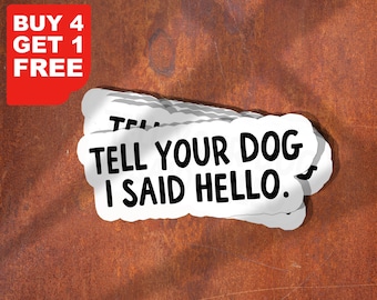 Op de afbeelding: Witte stickers met de zwarte tekst "TELL YOUR DOG I SAID HELLO", gestapeld op een roestbruine ondergrond. Een rode rechthoek in de linkerbovenhoek leest "BUY 4 GET 1 FREE" in wit.