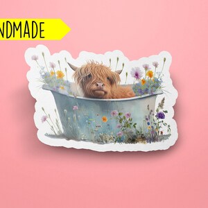 Peut inclure: Un autocollant fait main repr&eacute;sentant une vache Highland brune et blanche prenant un bain dans une baignoire bleue entour&eacute;e de fleurs sauvages color&eacute;es.