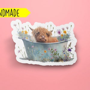 May include: A handmade sticker featuring a brown and white Highland cow taking a bath in a blue tub surrounded by colorful wildflowers.