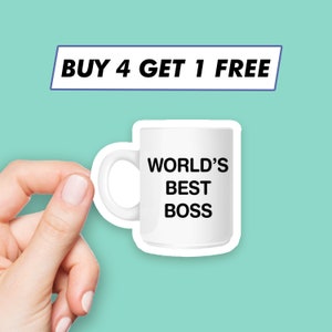 以下が含まれることがあります： 白いセラミック製のマグカップに黒い文字で「WORLD'S BEST BOSS」と書かれています。マグカップは手で持たれています。マグカップの上には青いバナーがあり、「BUY 4 GET 1 FREE」と書かれています。