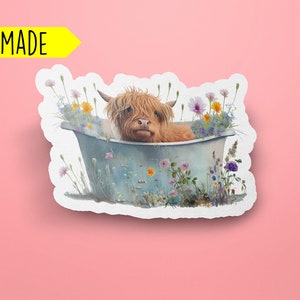 May include: A handmade sticker featuring a Highland cow taking a bath in a tub filled with wildflowers. The cow is brown and white with a fluffy coat. The tub is blue and white with a rustic look. The flowers are a variety of colors, including pink, purple, yellow, and white.