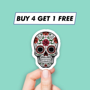Puede incluir: Una pegatina de calavera de azúcar blanca con detalles florales rojos y verdes. La pegatina tiene un fondo negro y se sostiene en una mano. El texto "BUY 4 GET 1 FREE" es visible sobre la pegatina.
