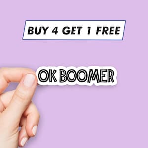 May include: A white sticker with black text that reads "OK BOOMER" with a white border. The sticker is being held by a hand against a light purple background. Above the sticker is a white rectangle with a blue border that reads "BUY 4 GET 1 FREE".