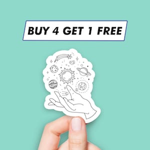 Op de afbeelding: Een zwart-witte sticker met een hand die een zon, planeten en een vallende ster vasthoudt. De sticker zegt "KOOP 4, KRIJG 1 GRATIS".