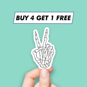 Puede incluir: Una pegatina blanca con un contorno negro que representa una mano esquelética haciendo el signo de la paz. La pegatina está sujeta por una mano sobre un fondo turquesa. El texto "BUY 4 GET 1 FREE" se muestra arriba.